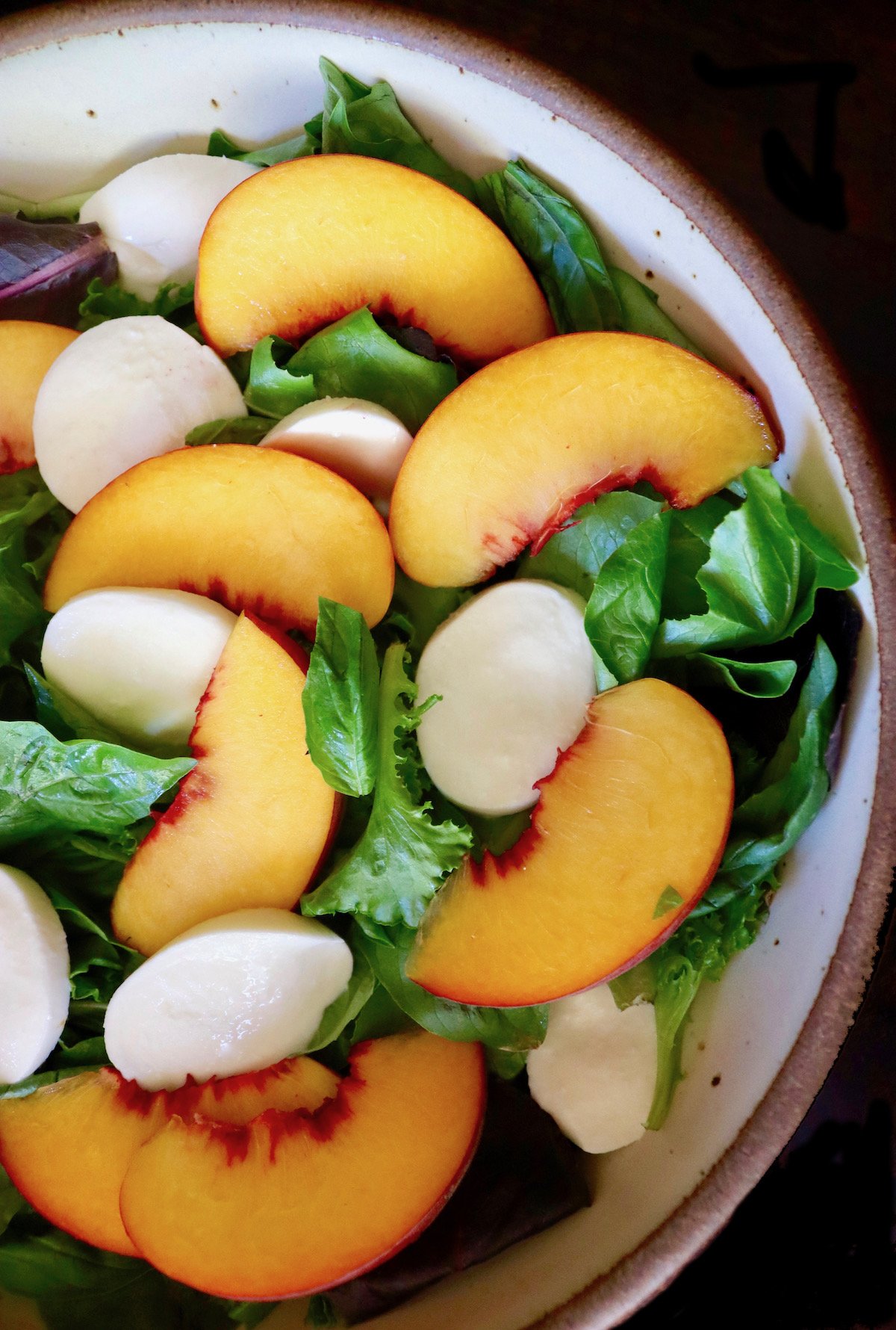 Sliced peaches, mozzarella and greens in a large cream-colored ceramic salad bowl.