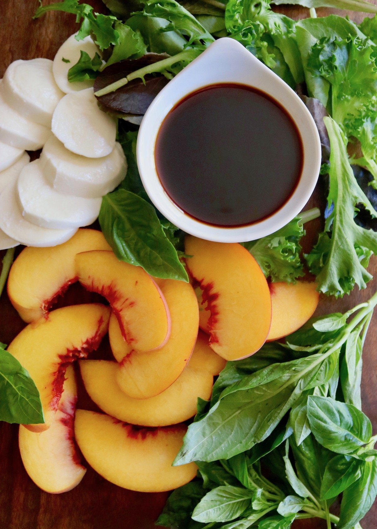 Sliced peaches, mozzarella, fresh basil leaves and lettuce, with a small white bowl of balsamic vinegar, all on a dark wood cutting board.
