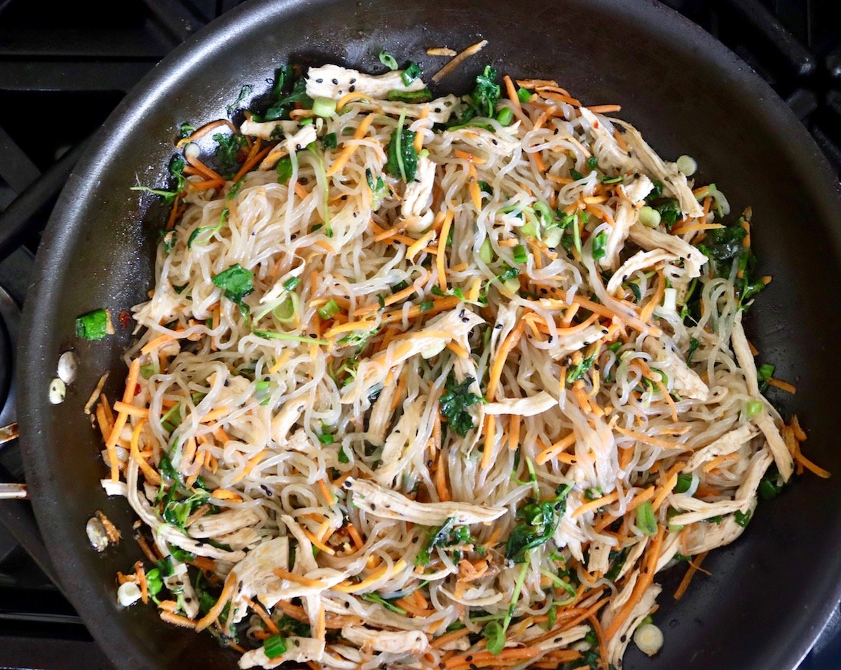 Large saute pan with clear noodles, shredded carrots and chicken with green herbs.