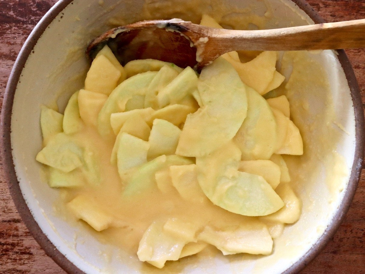 Egg mixture with apples slices and a wooden spoon, in a cream-colored ceramic bowl.