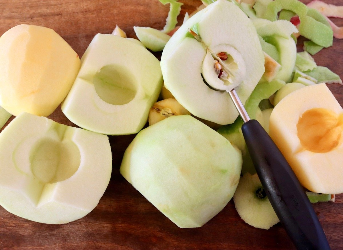 Several halved and peeled apples with a melon baller being used to core them.