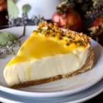 Close up of a slice of passion fruit cheesecake on white plate with whole passion fruits behind it.