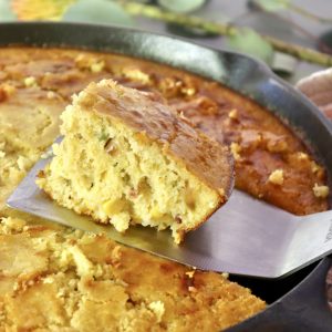 One square slice of cornbread with jalapeno and bacon being lifted from cast iron skillet with a metal spatula.