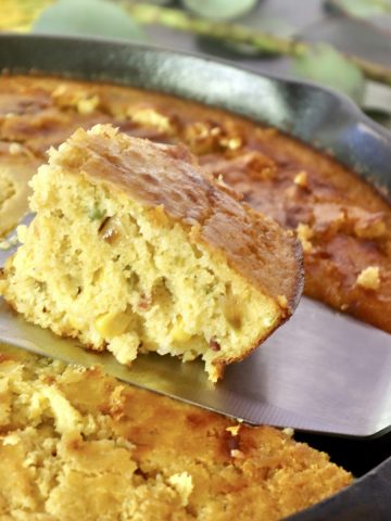 One square slice of cornbread with jalapeno and bacon being lifted from cast iron skillet with a metal spatula.