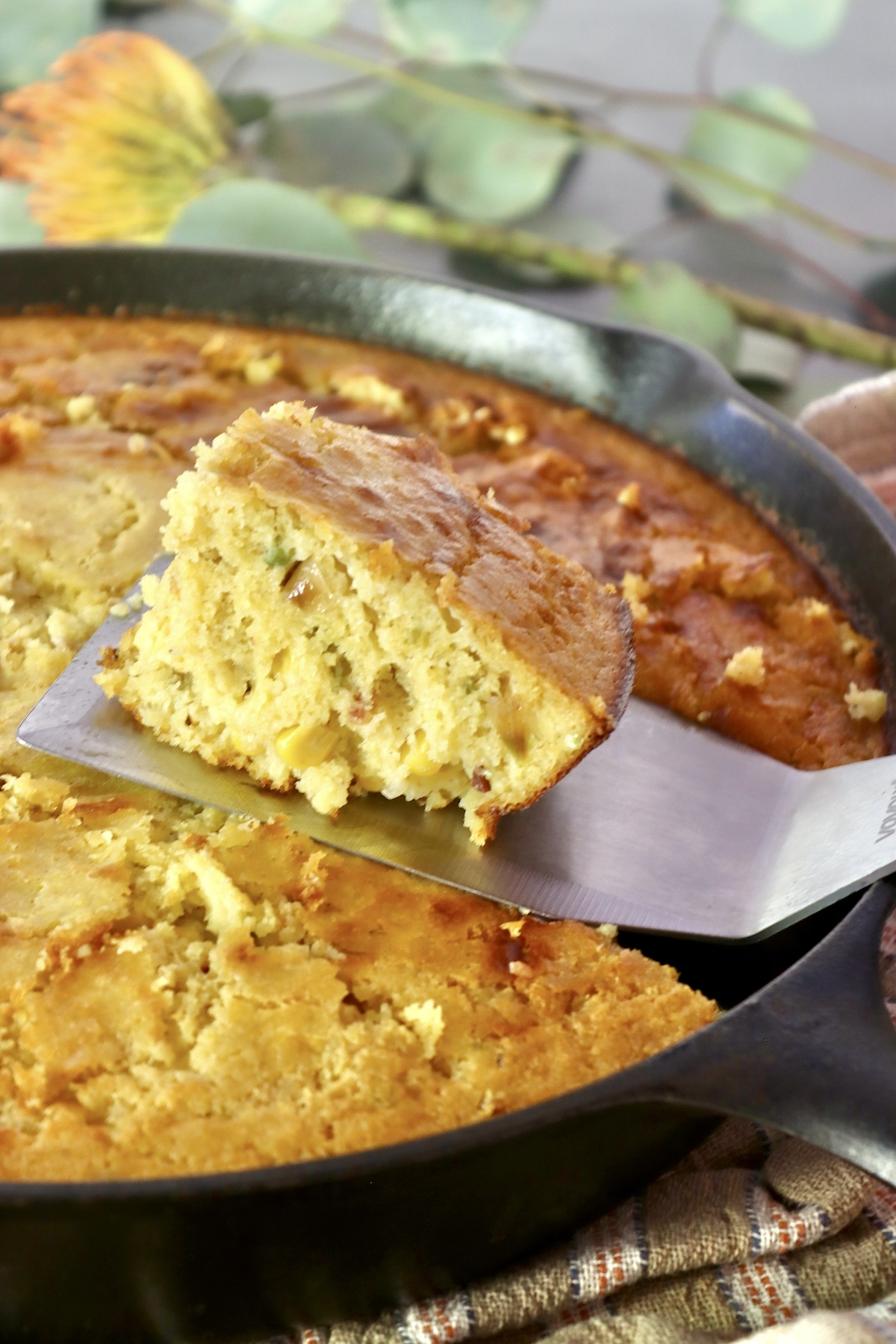 One square slice of cornbread with jalapeno and bacon being lifted from cast iron skillet with a metal spatula.