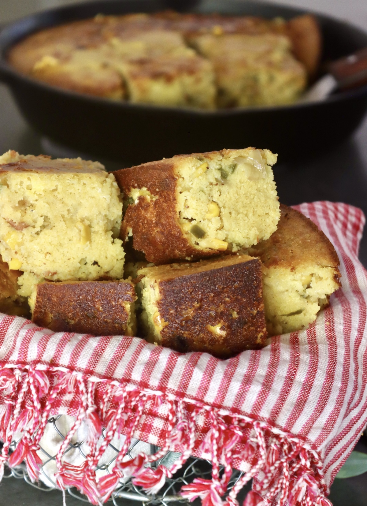 Several square slices of bacon jalapeno cornbread stacked on a red and white napkin, in front of cast iron skillet.