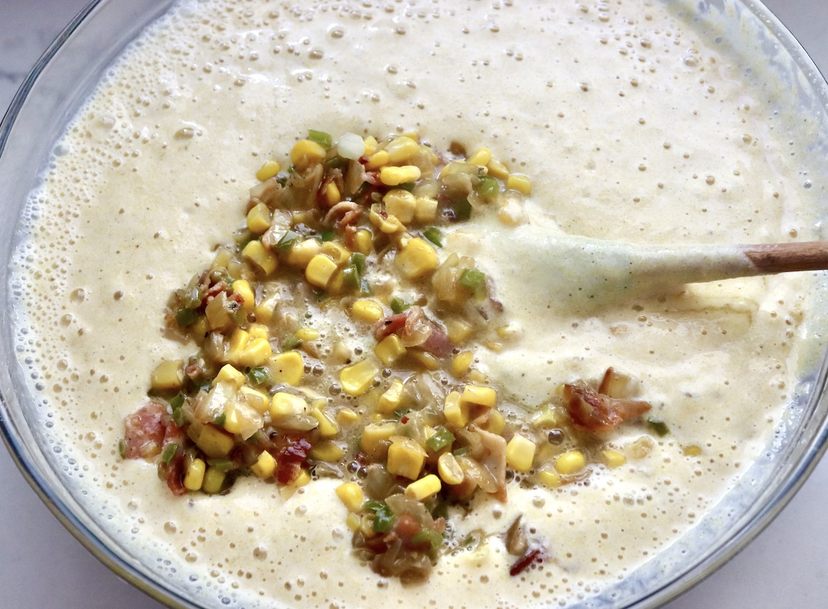 Cornbread batter in a mixing bowl with a wooden spoon an pile of corn kernels, crumbled bacon, diced onion and jalapeno.