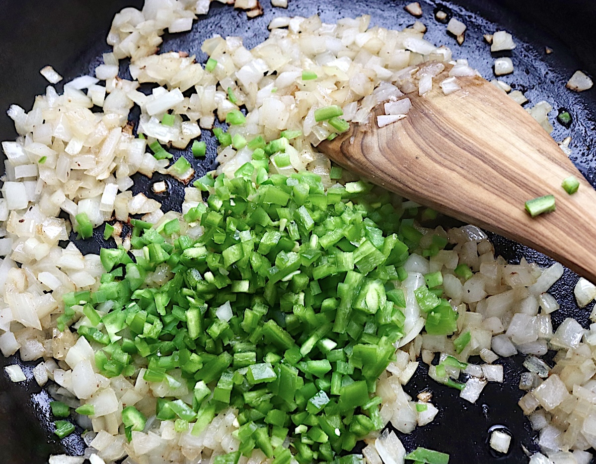 Diced onion and jalapeno pepper in a cast iron skillet with a wooden spatula.