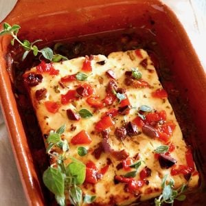 Baked Feta cheese with golden edges in a terra cotta baking dish with roasted peppers and olives.