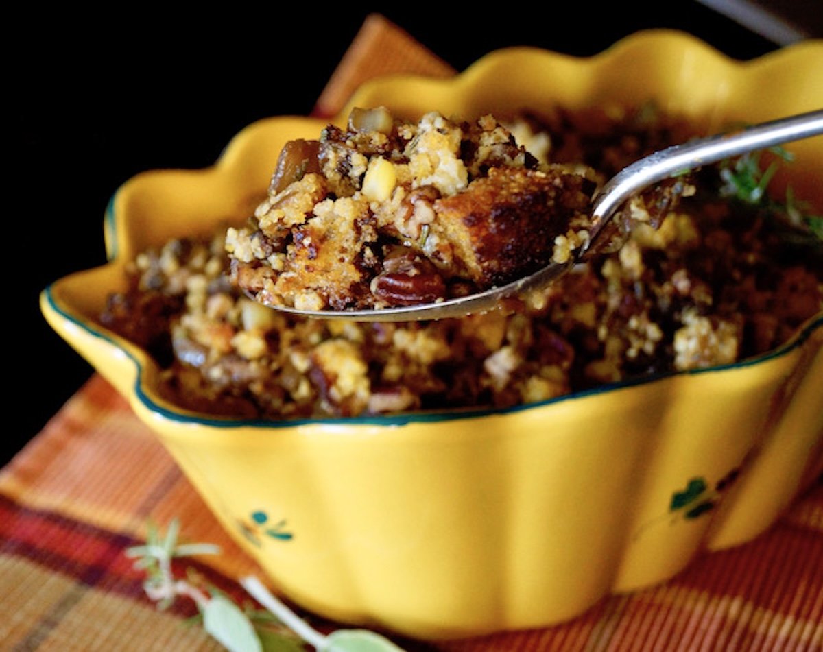 Pretty golden-yellow ceramic oval bowl full with corn bread stuffing, and a spoon holding up a large bite.