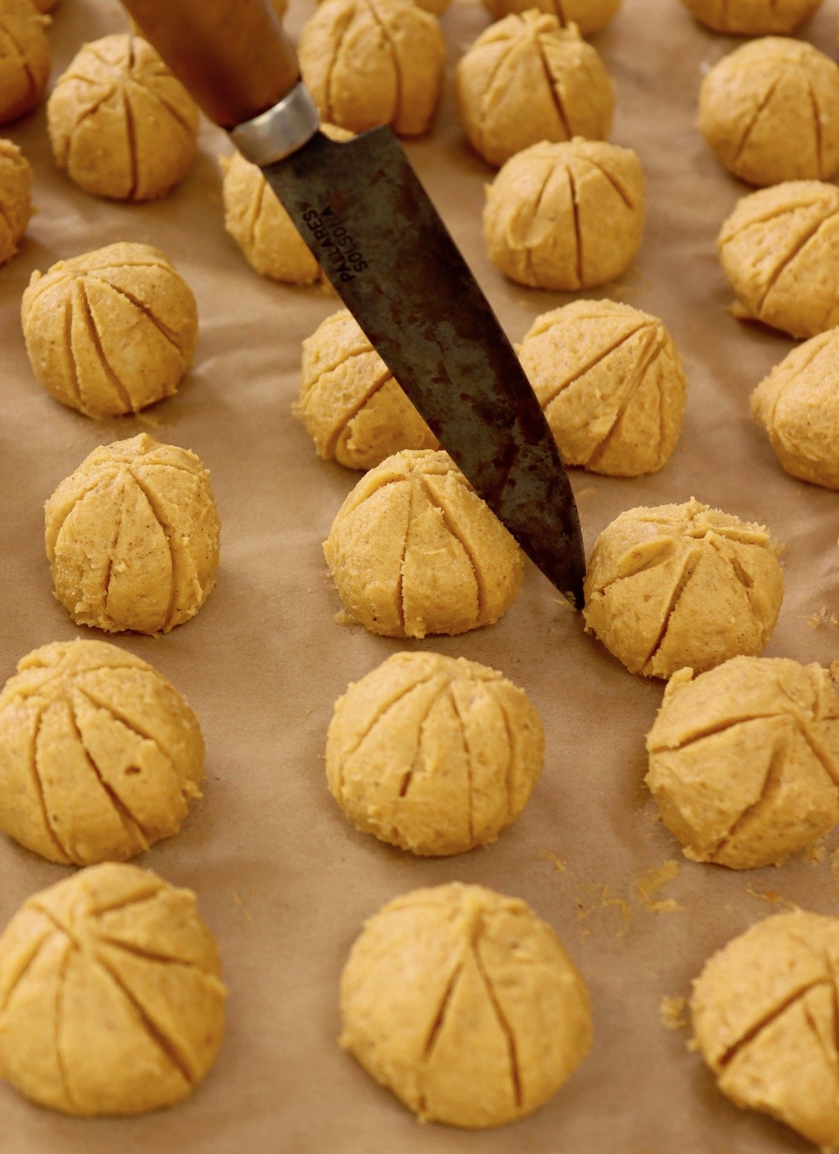 Over a dozen raw pumpkin cookie dough balls on parchment-lined baking sheet with parking knife to make pumpkin-like slits on the sides.