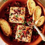 Terra cotta round baking dish filled with chopped olives and red peppers and two blocks of Feta cheese with toasts.