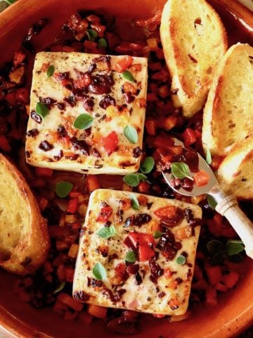 Terra cotta round baking dish filled with chopped olives and red peppers and two blocks of Feta cheese with toasts.