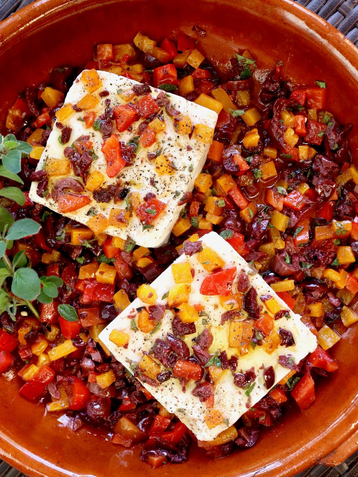 Two blocks of Feta cheese in a large terra cotta baking dish with chopped peppers and olives all around and on top of them, with sprig of oregano.