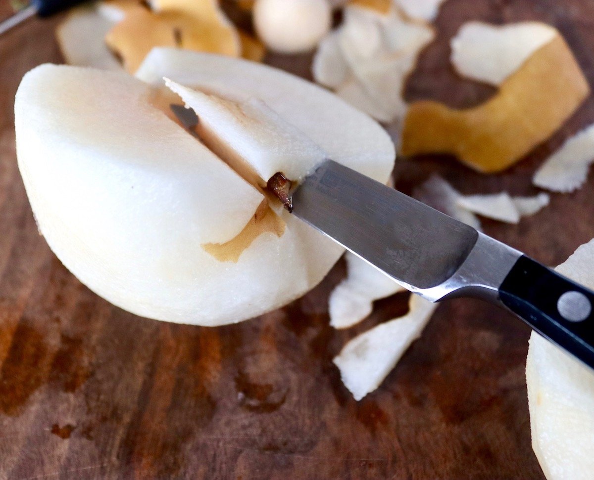 Half of a peeled Asian pear with a paring knife slicing bits of the root off.