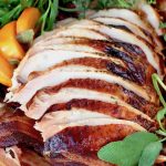 Sliced turkey breast with golden skin on cutting board with bacon, persimmon slices and fresh herbs.