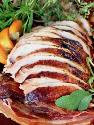 Sliced turkey breast with golden skin on cutting board with bacon, persimmon slices and fresh herbs.