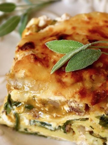 One slice of butternut squash and spinach lasagna on a white plate with fresh sage leaves.