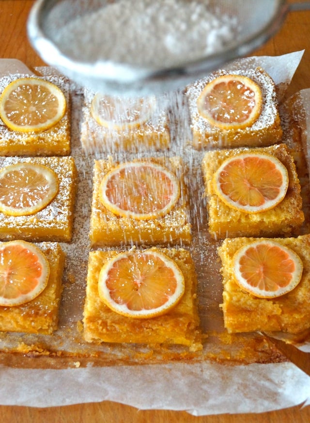 9 evenly spaced lemon bars that are square, on parchment paper with powdered sugar being sprinkled on them through mesh strainer.