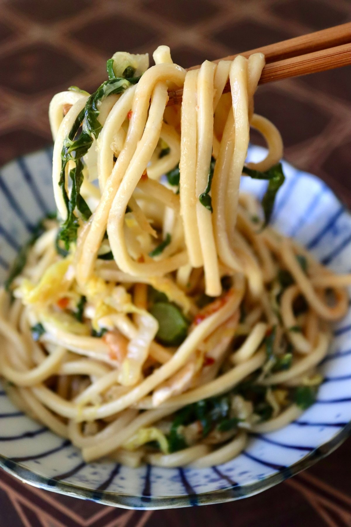 Serving of Chinese Longevity Noodles with Gai Lan on ceramic blue and white plate with a bite held up in chopsticks.
