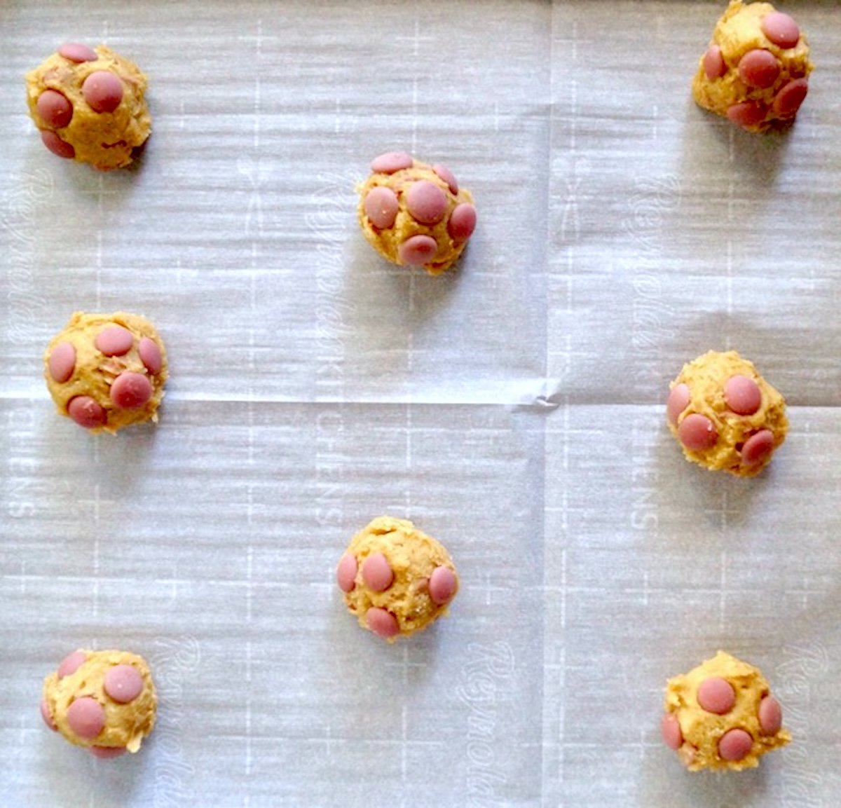 Several balls of chocolate chip cookie dough with pink Ruby chocolate chips, on white parchment paper.