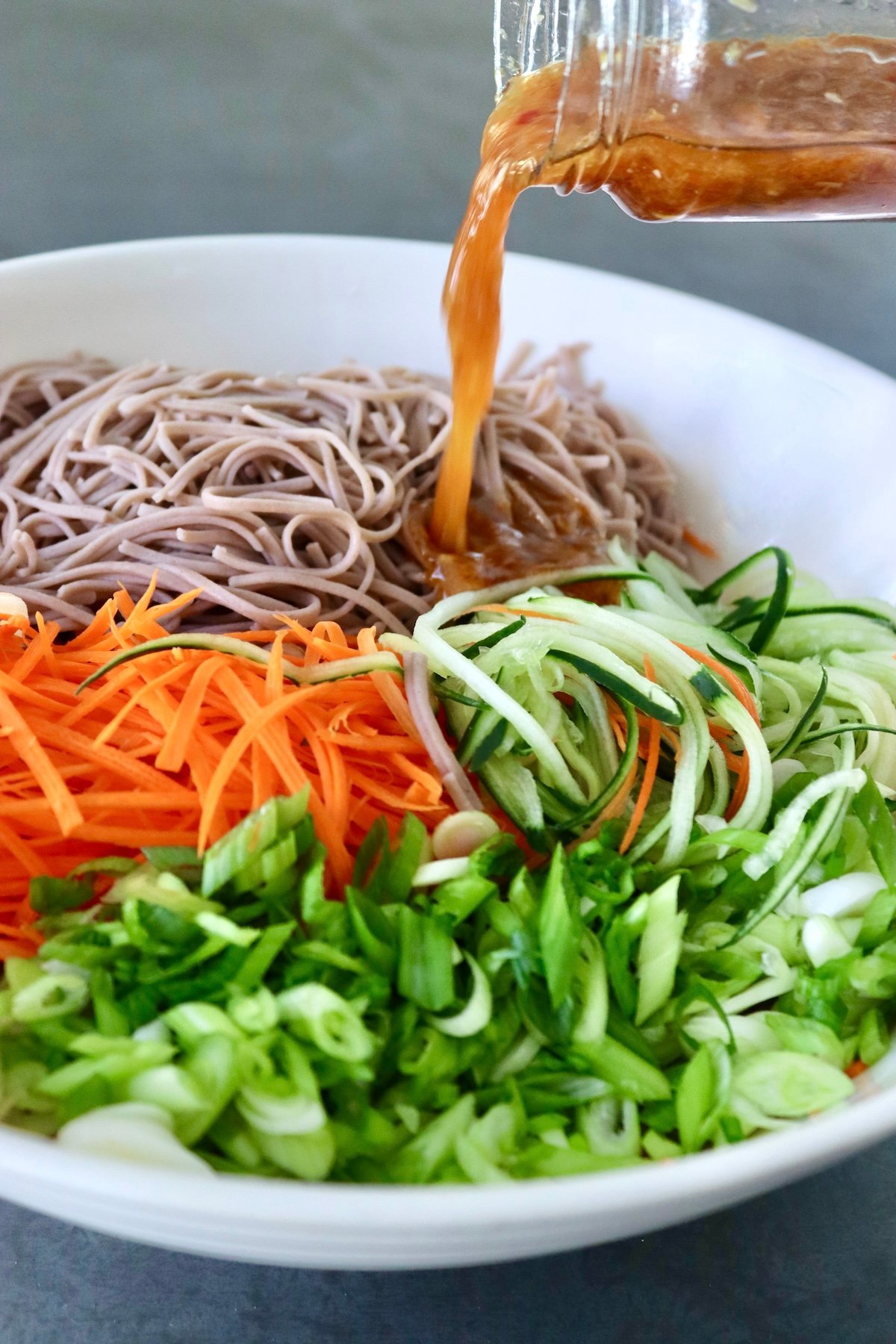White bowl full of soba noodles, carrots, cucumber  -- all shaped like noodles- and chopped scallions, with dressing being drizzled on top.