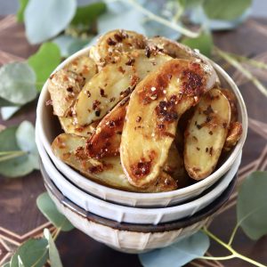 Three stacked cream-colored bowls with crispy fingerling potatoes filling the top one, surrounded by eucalyptus leaves.