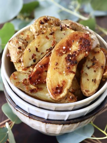 Three stacked cream-colored bowls with crispy fingerling potatoes filling the top one, surrounded by eucalyptus leaves.