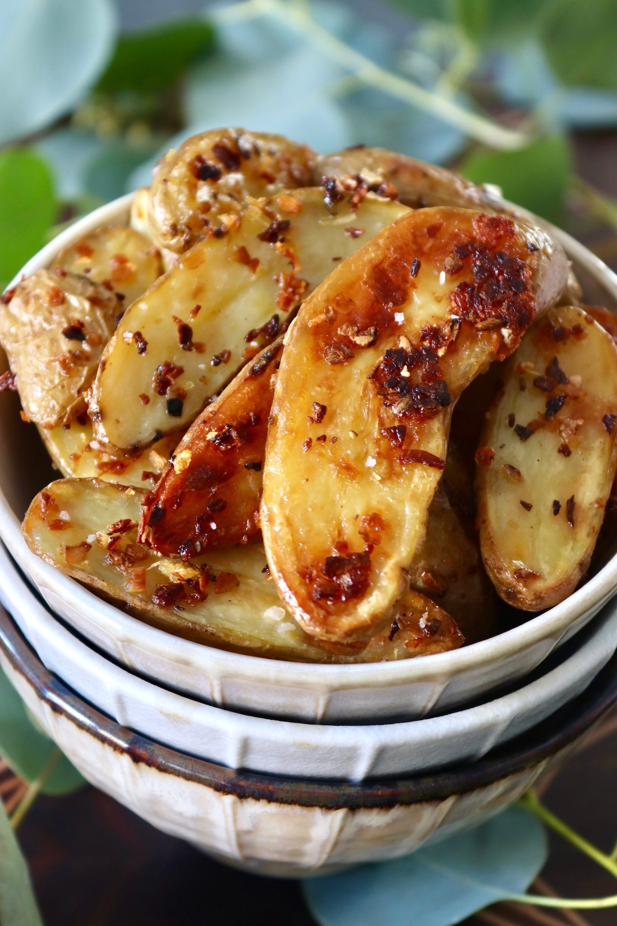 Close up of 3 stacked cream-colored bowls with crispy fingerling potatoes filling the top one, surrounded by eucalyptus leaves.