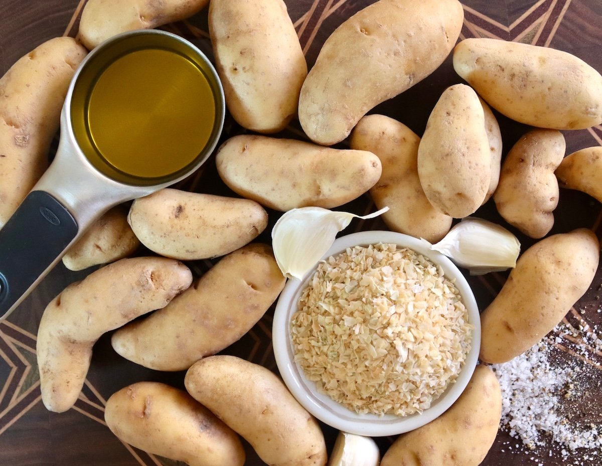 Many fingerling potatoes on a cutting board with a tiny bowl of dried minced onion, garlic cloves and olive oil in a measuring cup.