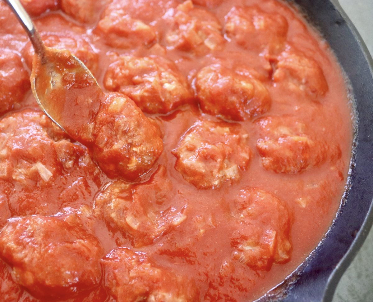 Cast iron skillet filled with tomato sriracha sauce and over a dozen meatballs, with a teaspoon flipping one of them.