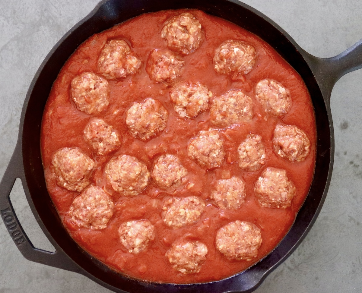 Cast iron skillet filled with tomato sriracha sauce and over a dozen raw meatballs.
