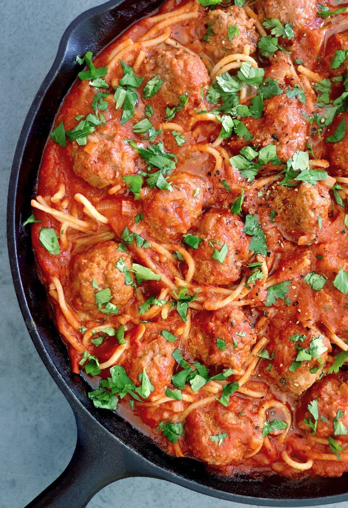 Close up of a cast iron skillet full with spaghetti and meatballs with Sriracha sauce and sprinkled cilantro.