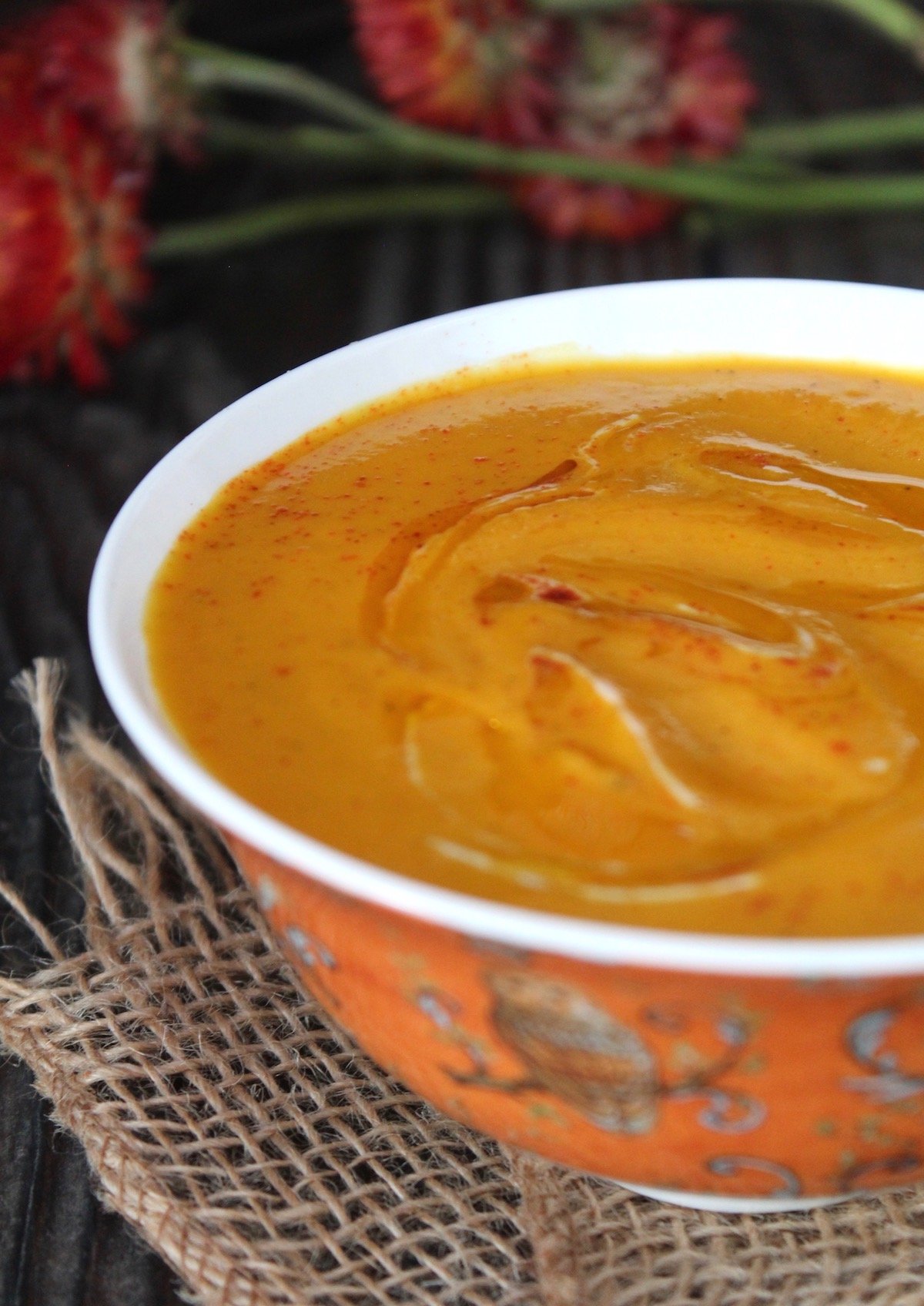 orange bowl with coconut spiced carrot soup and flowers behind it