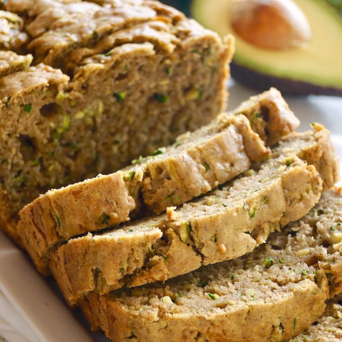 Sliced loaf of Avocado Zucchini Bread on a white plate with half of an avocado behind it, with pit.