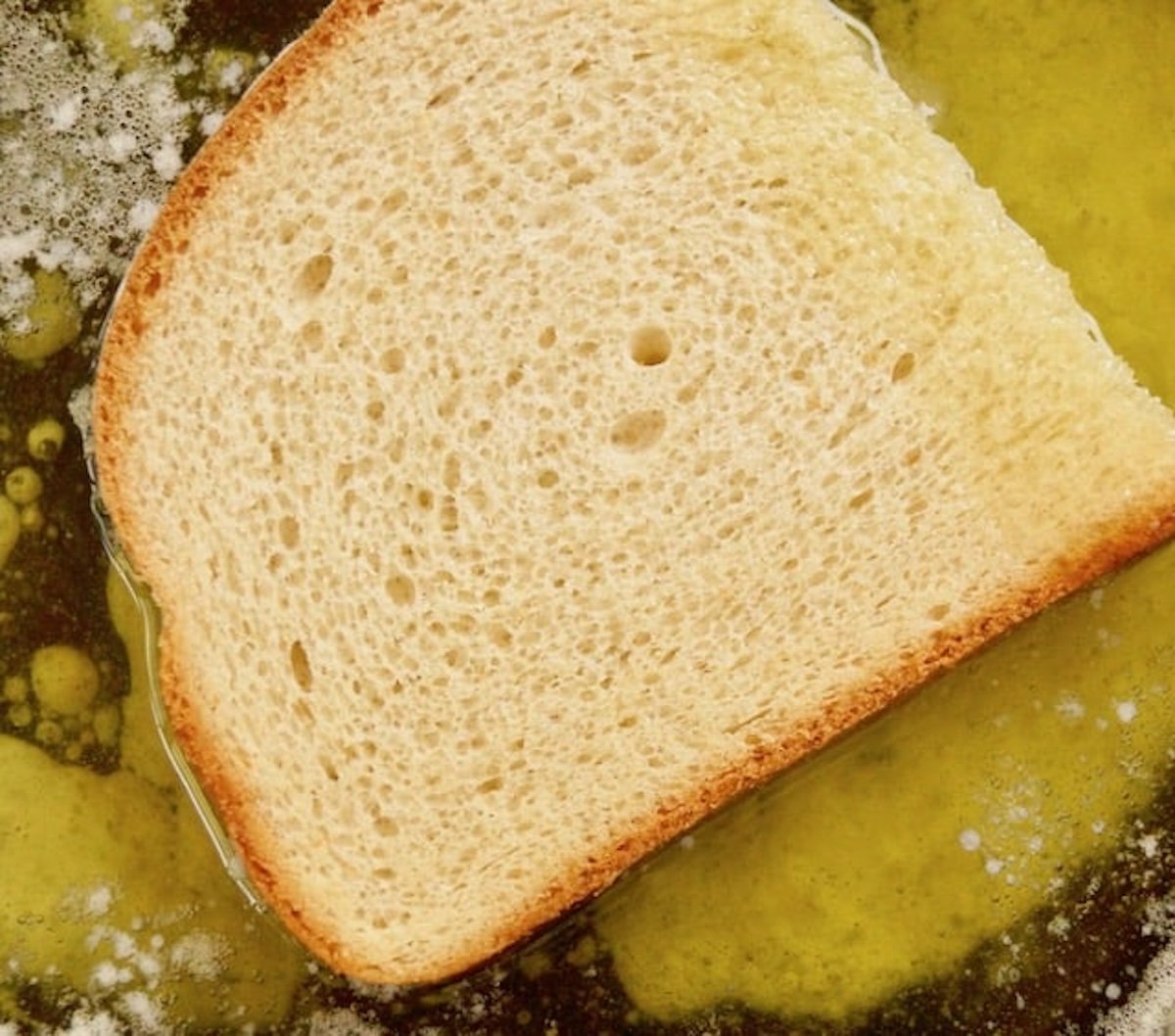 One slice of egg bead in a pan with lots of melted butter and oil beneath it.