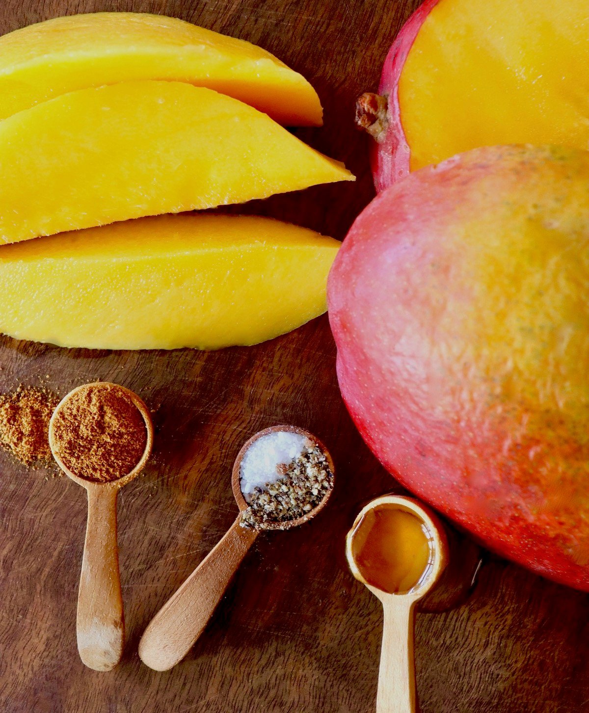 Sliced mango with half a red mango and three tiny wooden spoons with spices and honey, on a wood board.