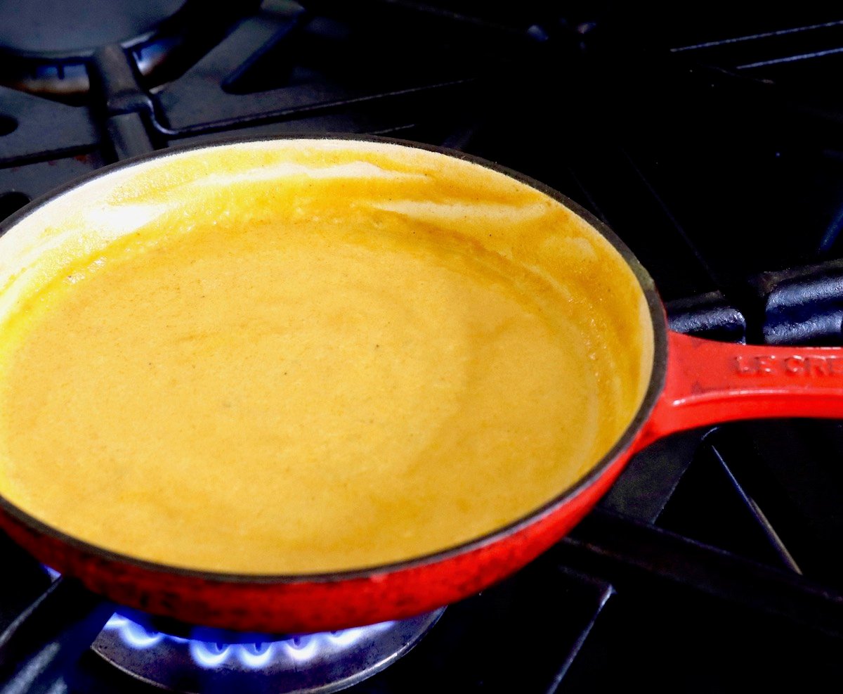 Small red sauce pan over a blue flame on a burner with mango coconut sauce.
