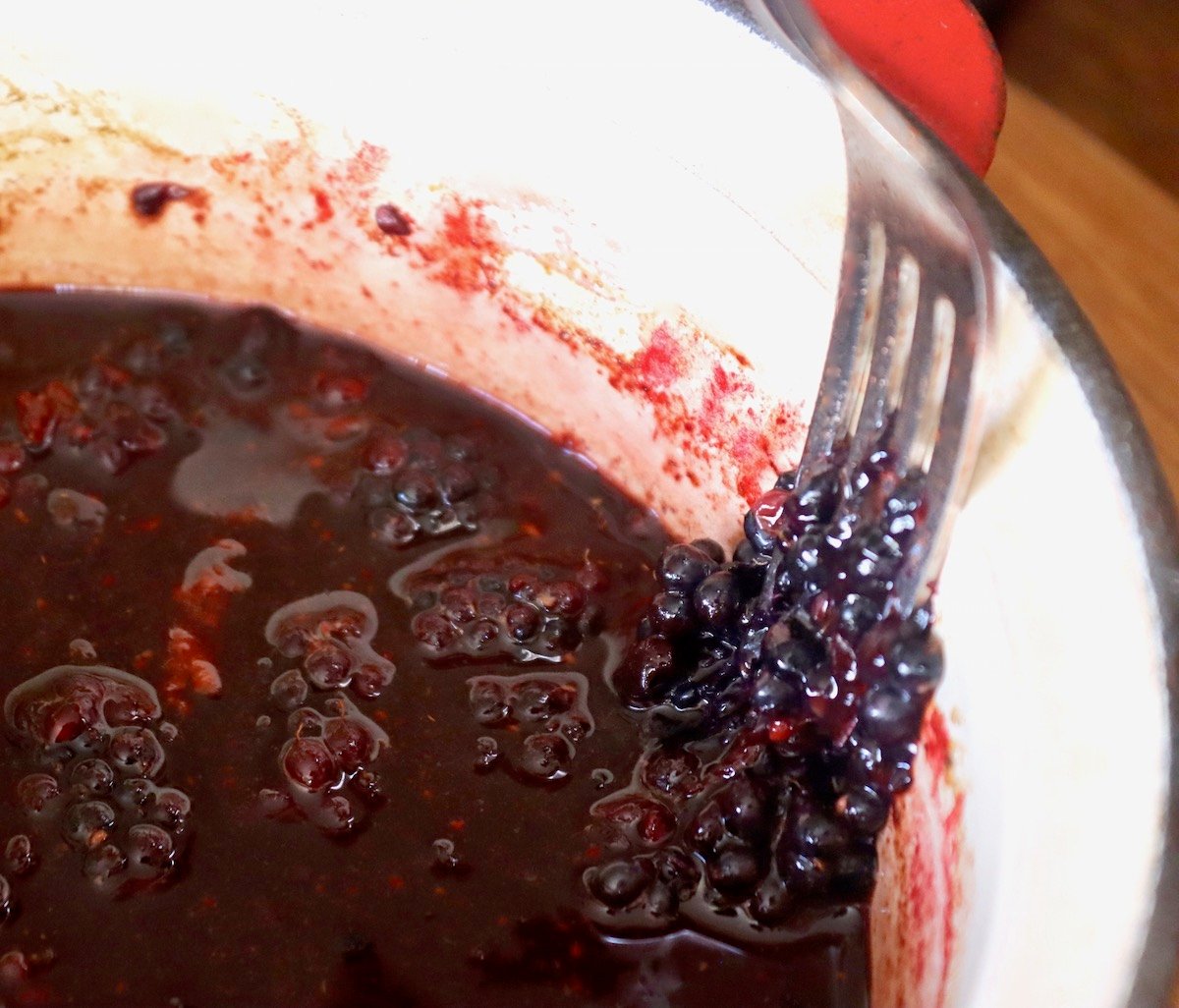 Small sauce pan with freshly cooked blackberry balsamic vinaigrette with a fork macerating the blackberries on the inside of the pan.