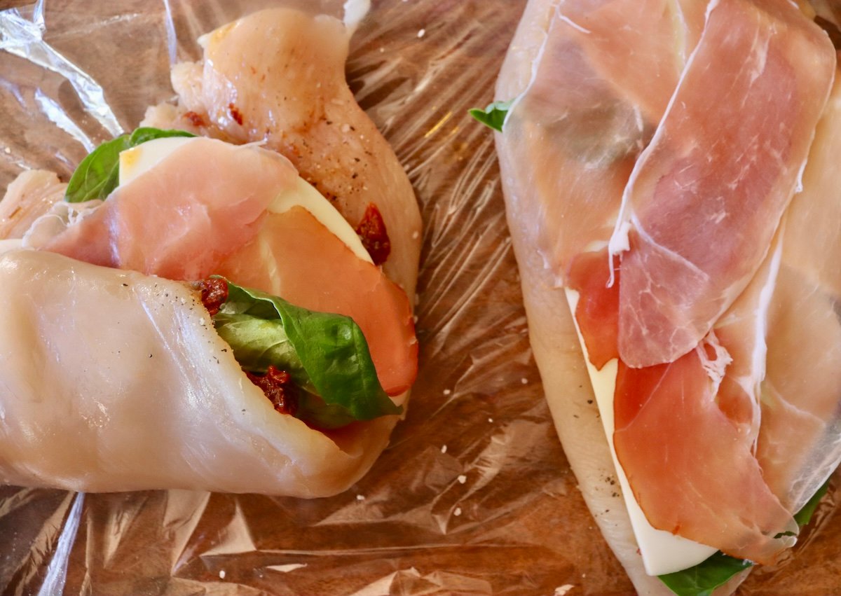 2 pounded chicken breasts with a layer of chopped sun-dried tomatoes, basil leaves, sliced provolone cheese and prosciutto slices on top, and one halfway rolled.