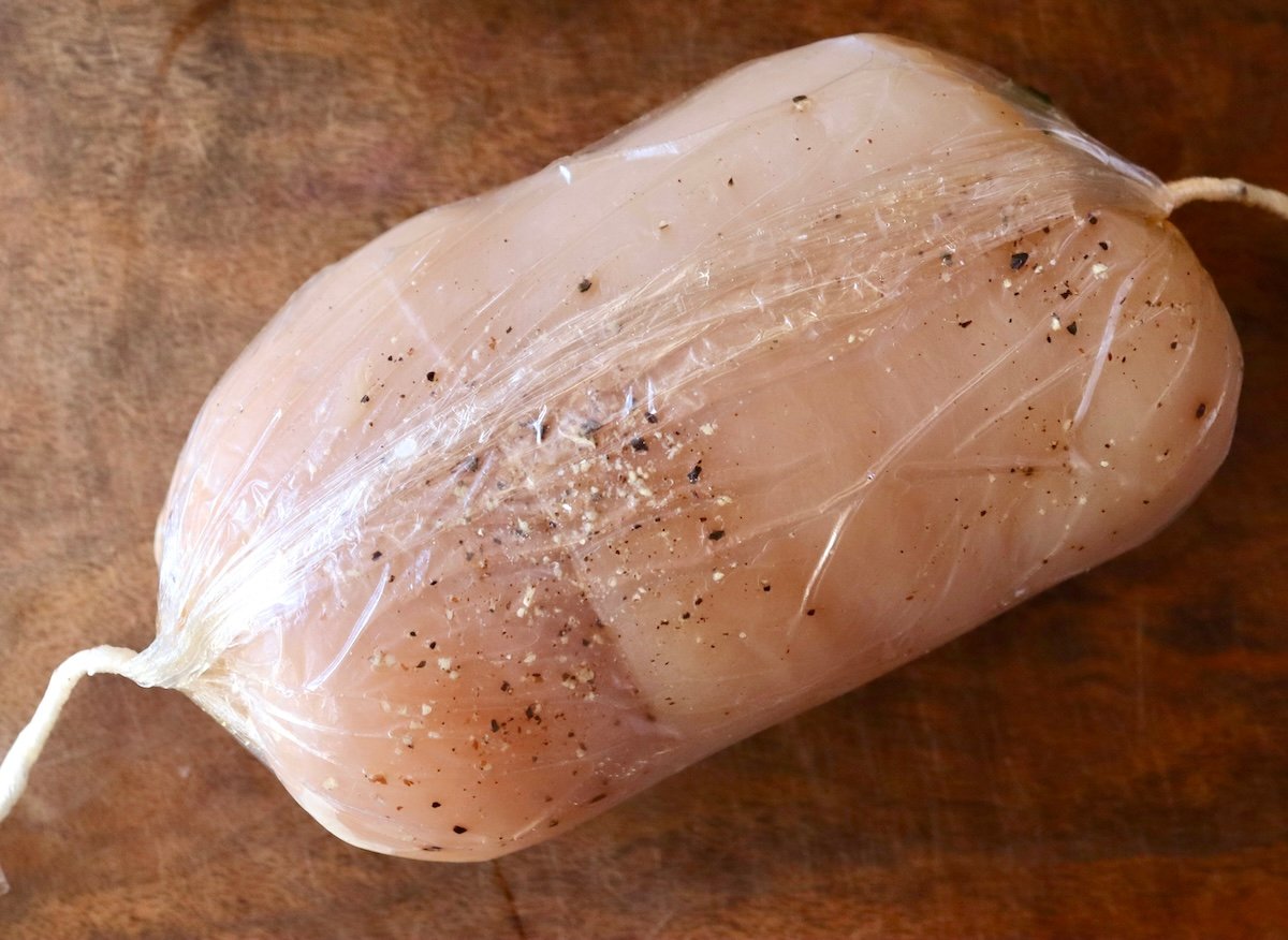 Raw chicken roulade with salt and pepper sprinkled on top, wrapped tightly in plastic wrap.