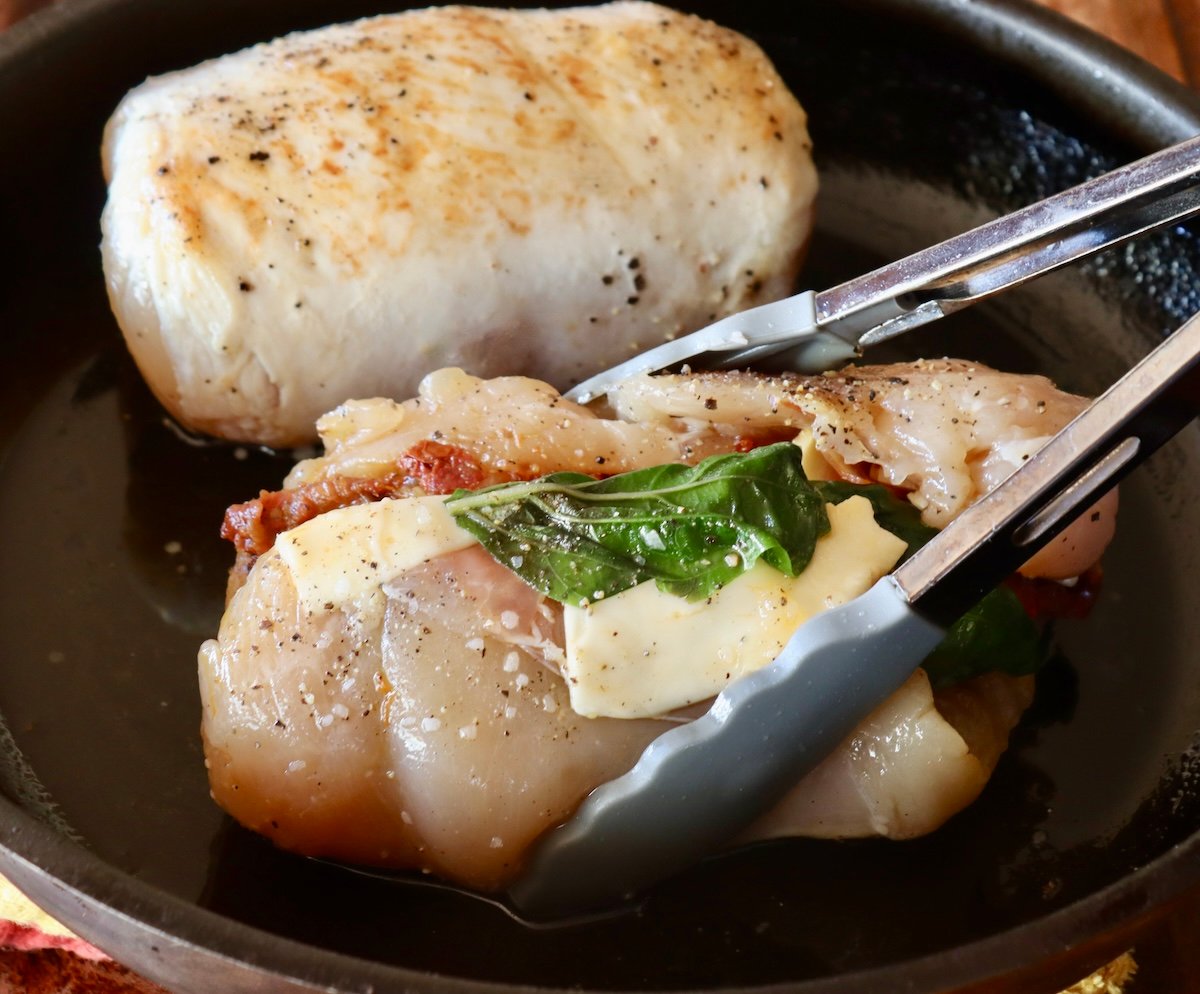 Two chicken roulades in a pan, one halfway cooked with tongs.