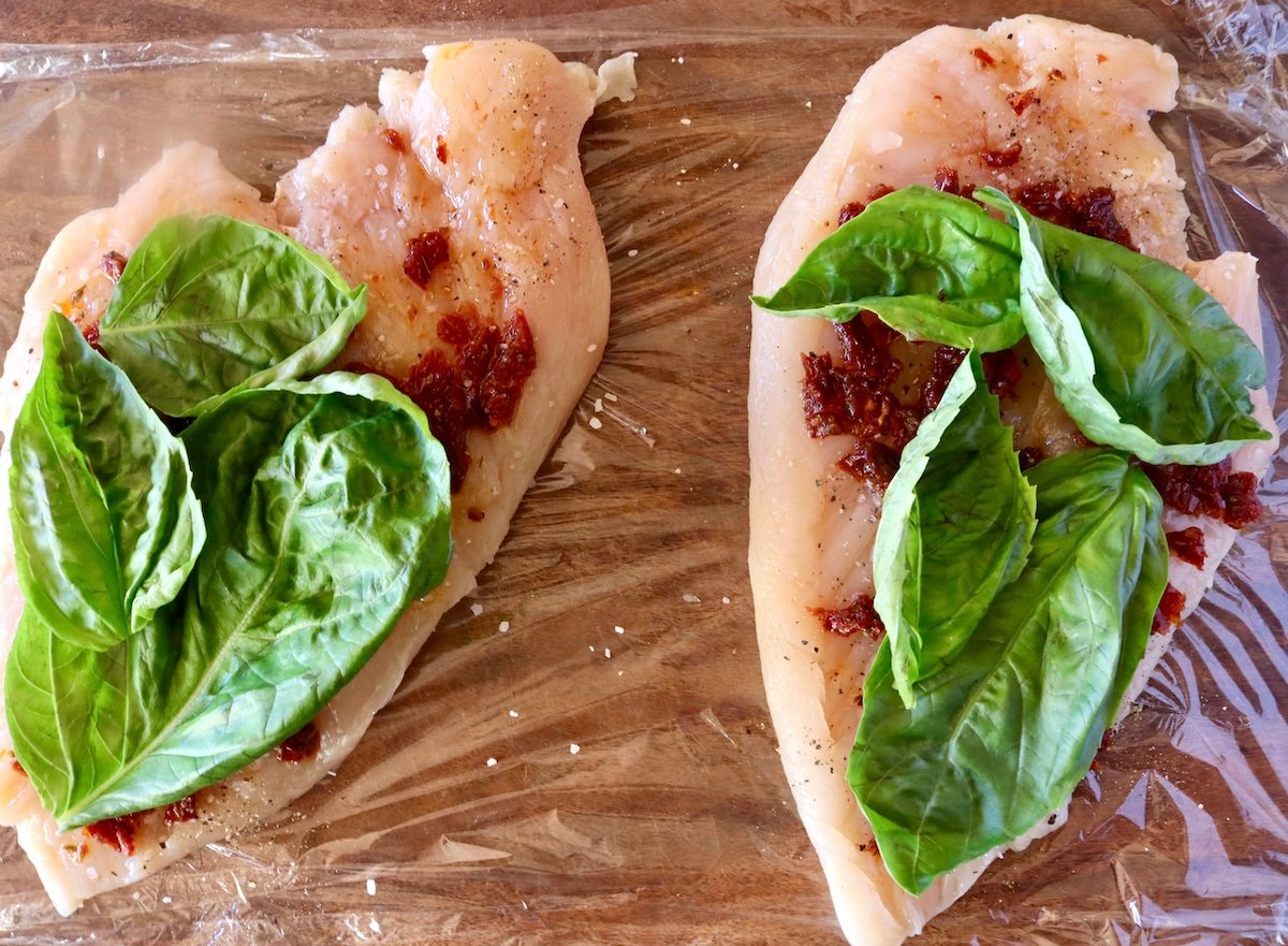 2 pounded chicken breasts with a layer of chopped sun-dried tomatoes and basil leaves on top.