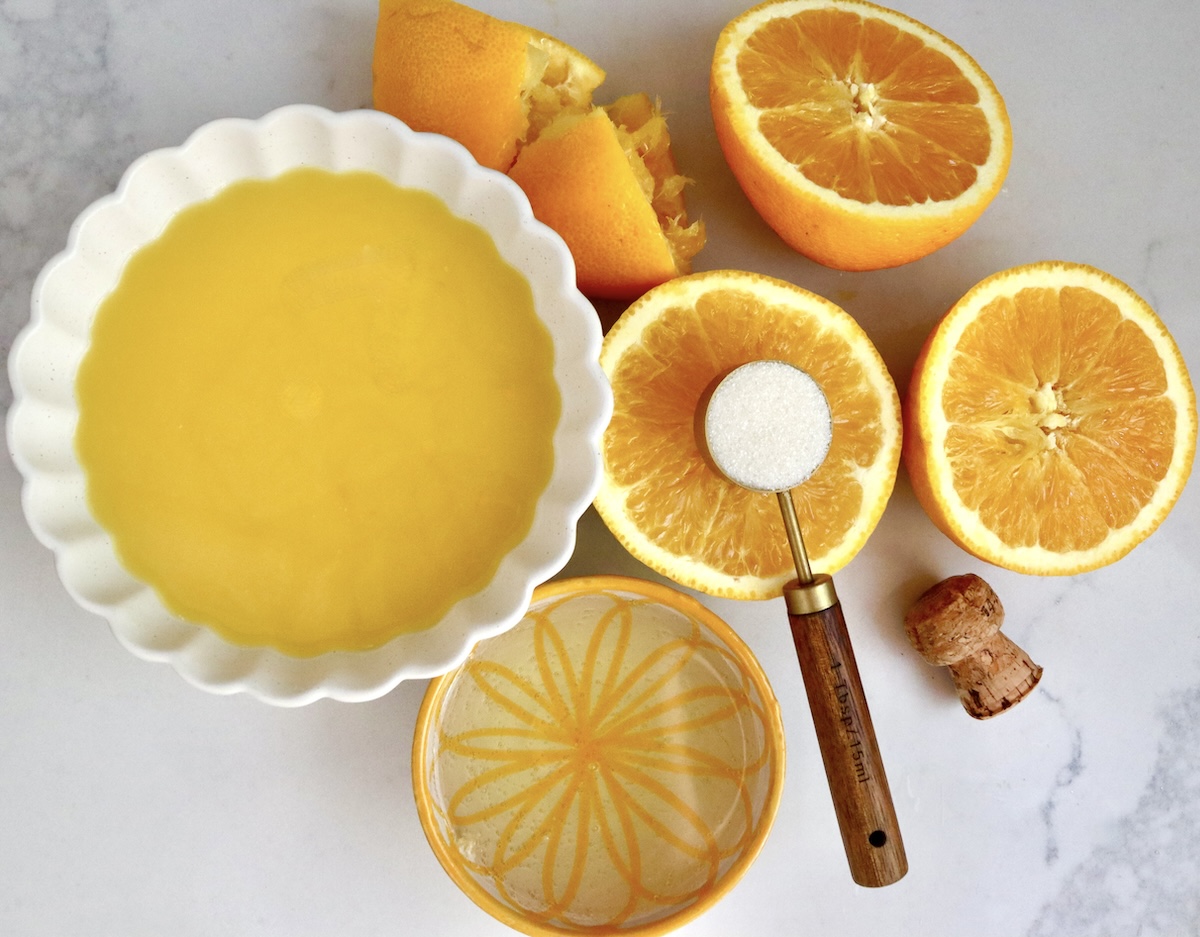 Frozen-Mimosa On white marble surface, there's a white crimp-edged bowl with orange juice, a few orange halves, tablespoon of sugar and a yellow and cream ceramic painted bowl with champagne.
