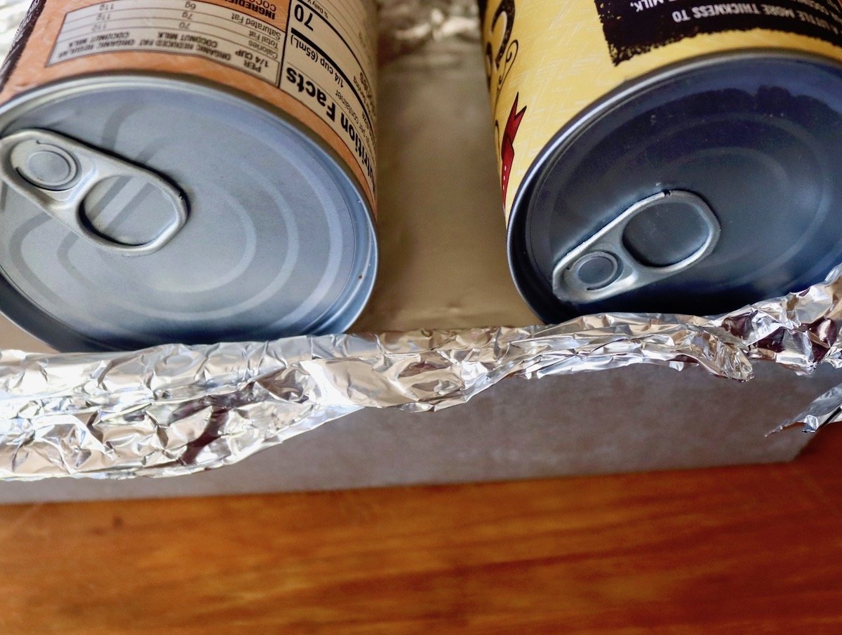 Foil-covered loaf pan with cans of food on top.