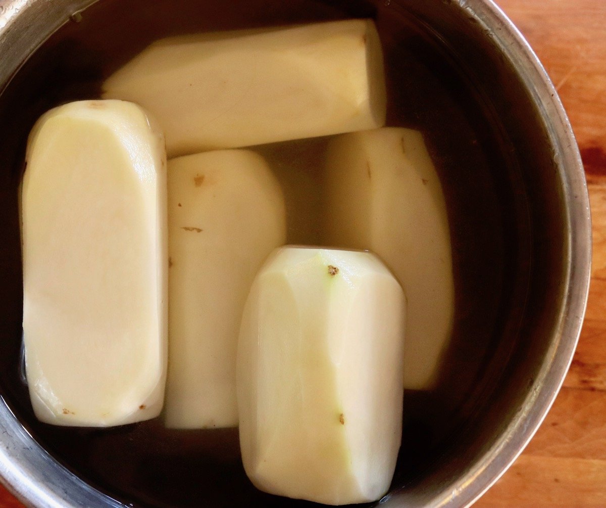 Potatoes peeled and cut into retangular shapes, in bowl of water.