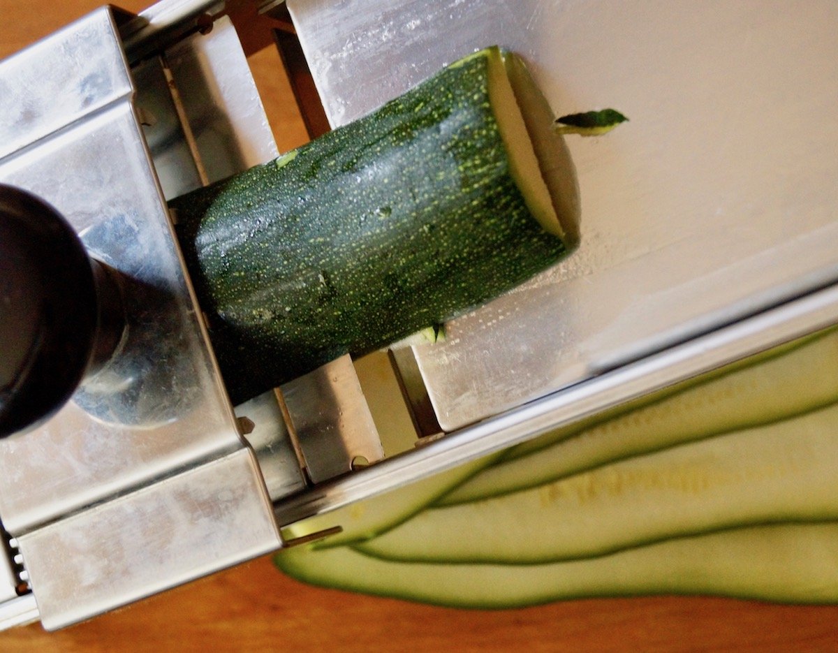 Zucchini being sliced into thin strips on a mandoline.