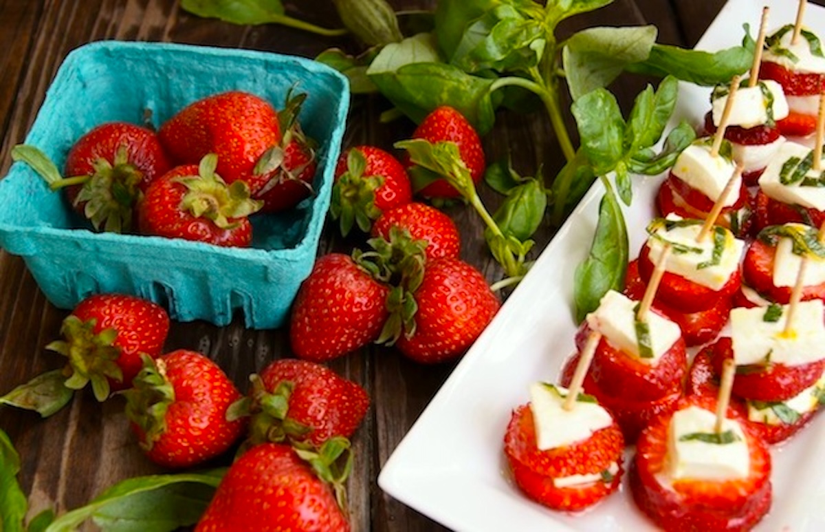 Strawberry-Caprese-with-Lemon-and-Basil Turquoise cardboard strawberry box with strawberries in and around it, with basil leaves next to it, and a white plate with strawberry caprese with toothpicks in them.