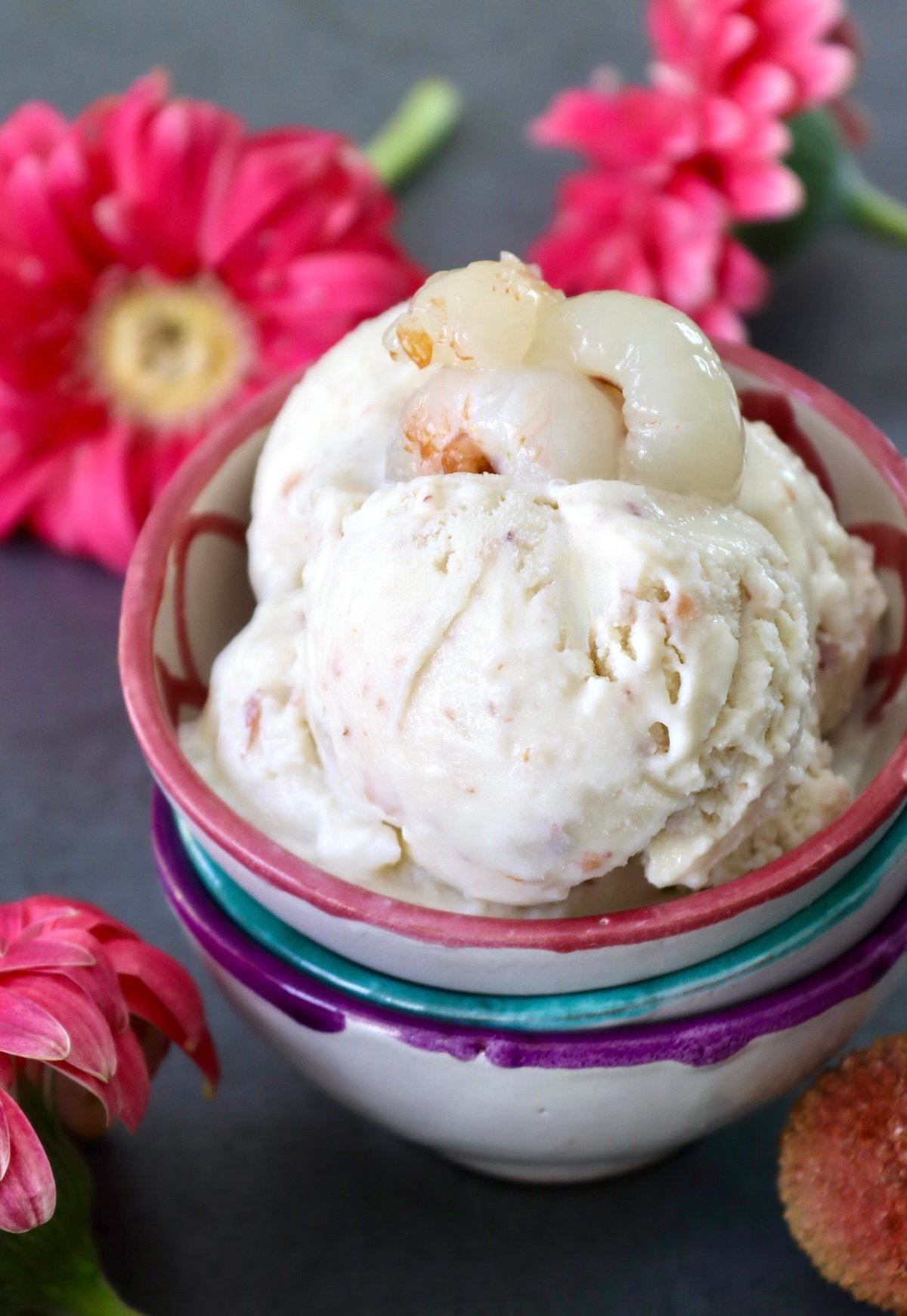 Lychee-Ice-Cream-Recipe Pink rimmed ceramic bowl with a generous few scoops of lychee ice cream, with a few fresh lychee on top, andbright pink Gerbera daisies in the background.