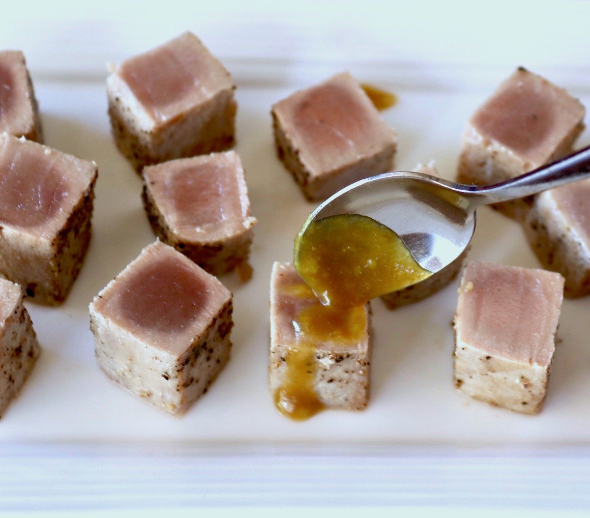Several small cubes of seared ahi tuna on a white plate with wasabi glaze being drizzled over one.